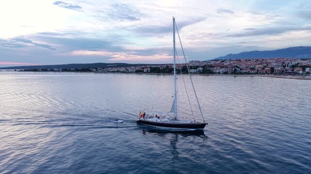 Aerial view of a large sailing yacht cruising across calm Adriatic waters near Novalja, Croatia, with the coastal town and dramatic rocky Velebit mountains in the background.