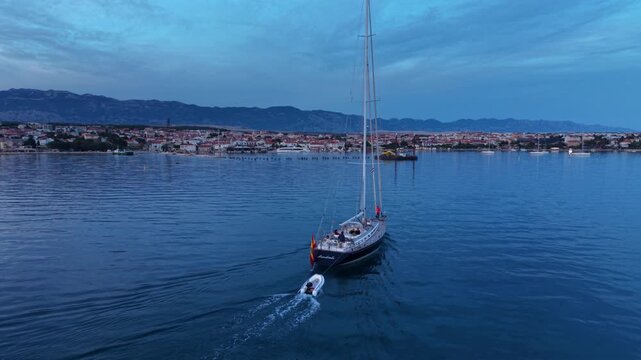 Aerial view of a large sailing yacht cruising across calm Adriatic waters near Novalja, Croatia, with the coastal town and dramatic rocky Velebit mountains in the background.