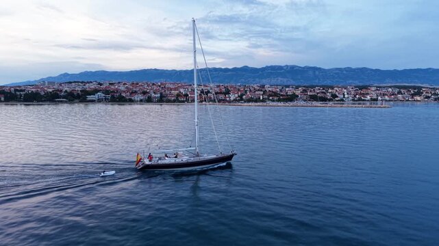 Aerial view of a large sailing yacht cruising across calm Adriatic waters near Novalja, Croatia, with the coastal town and dramatic rocky Velebit mountains in the background.