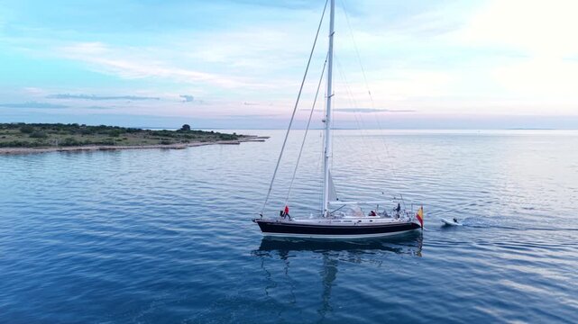 Aerial view of a large sailing yacht cruising across calm Adriatic waters near Novalja, Croatia, with the coastal town and dramatic rocky Velebit mountains in the background.