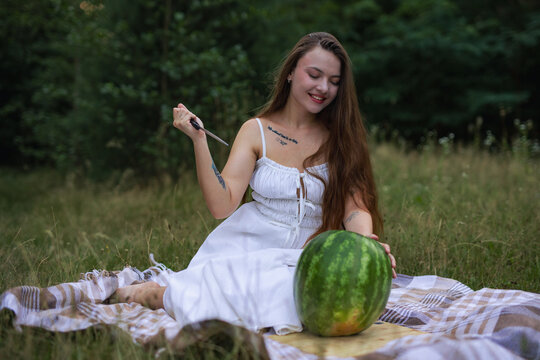 Smiling young woman holding knife to cut large watermelon on picnic blanket