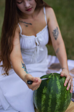 Young woman looking down while cutting watermelon on picnic blanket