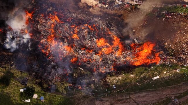 Aerial drone view of a major landfill fire with intense flames and thick black smoke rising from burning waste, as firefighters and emergency vehicles respond beside a recycling facility.