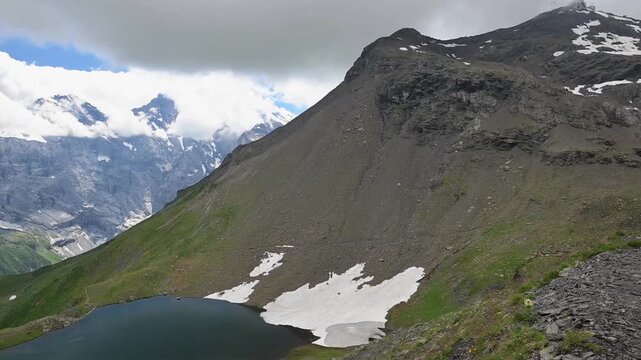 Turquoise mountain lake surrounded by rugged green slopes, lingering snowfields and dramatic Swiss Alps under cloudy skies.