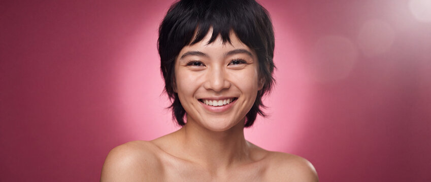 Asian woman smiling cheerfully with short hair against a vibrant pink studio backdrop, showcasing glowing natural skin for skincare, beauty, and wellness advertising campaign