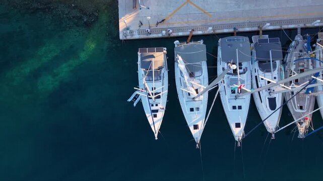 Top-down aerial view of several sailing yachts moored side by side at a marina pier, with their masts creating strong geometric lines above deep blue transparent water near a rocky shoreline.
