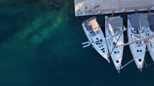 Top-down aerial view of several sailing yachts moored side by side at a marina pier, with their masts creating strong geometric lines above deep blue transparent water near a rocky shoreline.