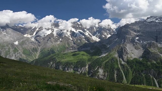 Dramatic Swiss Alpine valley with snow-covered peaks, green mountain slopes, rocky cliffs, and summer clouds near Birg.