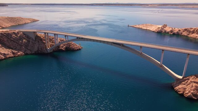 Aerial view of the Pag Bridge in Croatia spanning the narrow Ljubačka vrata sea channel, with its elegant concrete arch connecting Pag Island to the mainland against the rugged Velebit Mountains and d