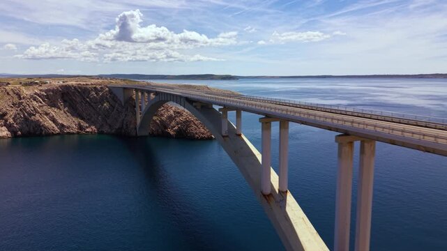 Aerial views of the Pag Bridge in Croatia carrying road traffic across a narrow Adriatic Sea channel, with the concrete arch structure spanning deep blue water between the barren rocky landscapes of P
