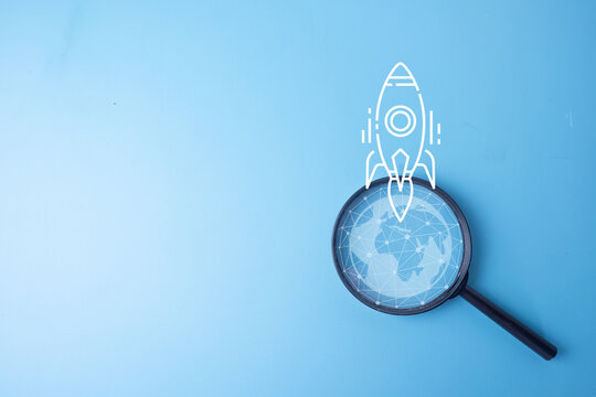 Global business startup and innovation concept. Magnifying glass focusing on a digital wireframe globe with a white rocket ship launching on a blue background with copy space.
