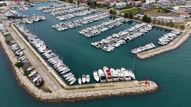 Aerial view of a large Mediterranean marina packed with yachts and sailboats, with long stone breakwaters, multiple piers and dense rows of moored vessels beside a coastal residential area.