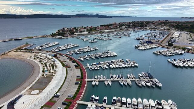 High aerial view of a large Mediterranean marina filled with rows of yachts and sailboats, protected by a curved breakwater beside a coastal town and waterfront promenade on the Adriatic coast.