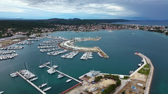 High aerial view of a large Mediterranean marina filled with rows of yachts and sailboats, protected by a curved breakwater beside a coastal town and waterfront promenade on the Adriatic coast.