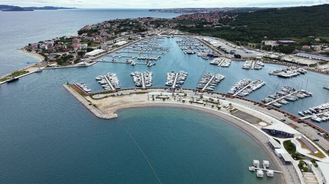 High aerial view of a large Mediterranean marina filled with rows of yachts and sailboats, protected by a curved breakwater beside a coastal town and waterfront promenade on the Adriatic coast.