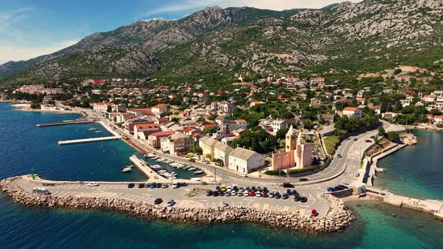 Top-down aerial view of a small historic church in Karlobag, Croatia, beside a curved coastal road with several motorcycles riding past, surrounded by parking areas and traditional red-roofed building