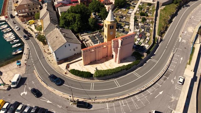 Top-down aerial view of a small historic church in Karlobag, Croatia, beside a curved coastal road with several motorcycles riding past, surrounded by parking areas and traditional red-roofed building