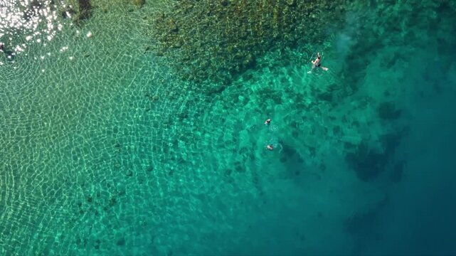 Top-down aerial view of swimmers floating and snorkeling in crystal-clear turquoise sea, with shallow rocky seabed patterns fading into deeper blue water along a pristine coastal area.