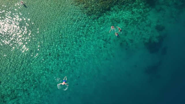 Top-down aerial view of swimmers floating and snorkeling in crystal-clear turquoise sea, with shallow rocky seabed patterns fading into deeper blue water along a pristine coastal area.