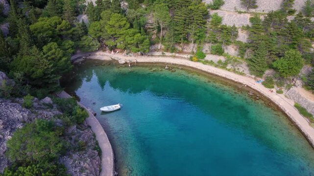 Aerial view of a secluded rocky cove with clear turquoise water, a narrow coastal path and dense pine forest along the shoreline in a peaceful Mediterranean landscape.