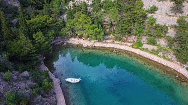 Aerial view of a secluded rocky cove with clear turquoise water, a narrow coastal path and dense pine forest along the shoreline in a peaceful Mediterranean landscape.
