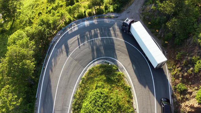 Top-down aerial drone view of a semi truck negotiating a tight hairpin bend on a mountain road surrounded by dense green forest, with cars passing through the sharp curve.