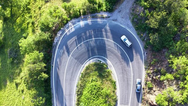 Top-down aerial drone view of a semi truck negotiating a tight hairpin bend on a mountain road surrounded by dense green forest, with cars passing through the sharp curve.