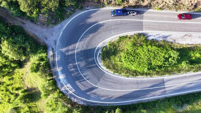 Top-down aerial drone view of a semi truck negotiating a tight hairpin bend on a mountain road surrounded by dense green forest, with cars passing through the sharp curve.