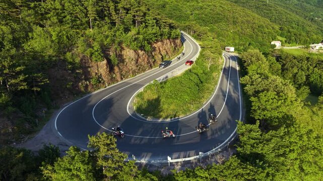 Aerial drone view of motorcyclists riding around a dramatic hairpin bend on a winding mountain road surrounded by dense green forest, with cars and a truck traveling nearby.