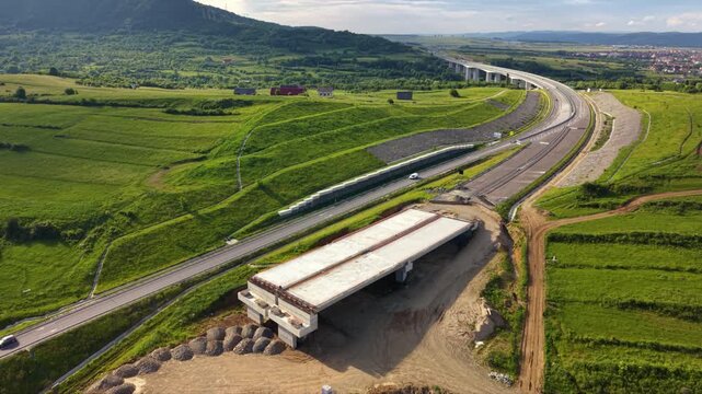 Aerial drone view of highway bridge construction across green rural fields. Concrete piers, cranes, earthworks and unfinished overpass sections show major transport infrastructure development project.