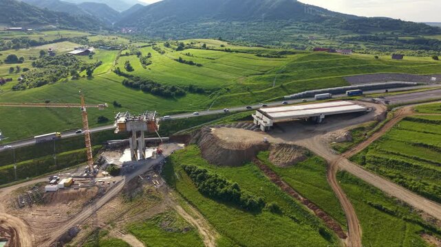 Aerial drone view of highway bridge construction across green rural fields. Concrete piers, cranes, earthworks and unfinished overpass sections show major transport infrastructure development project.