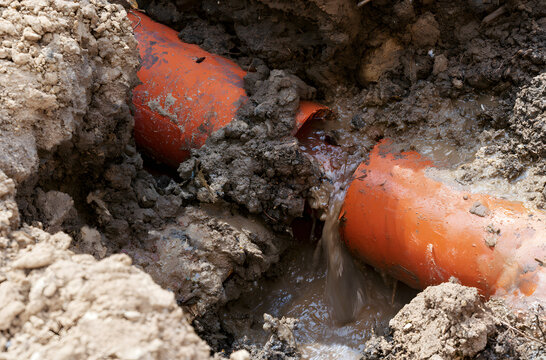 An orange, damaged underground pipe, which was leaking water and mud from an excavated trench, highlights plumbing mishaps, infrastructure destruction, and pipe repair projects