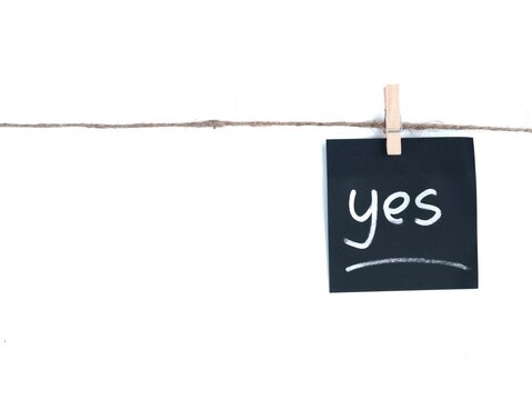 A single black square paper note with the white handwritten word yes is clipped by a wooden clothespin to a natural jute twine string. The background is pure white with empty space on the left