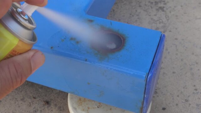 Close-up of a hand using an aerosol spray to exterminate a wasp nest built inside a rusty blue metal structure. Pest control concept