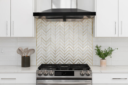 A white kitchen detail with a stainless steel range hood over a has range, a gold and marble chevron backsplash, subway tiles, and a marble countertop.