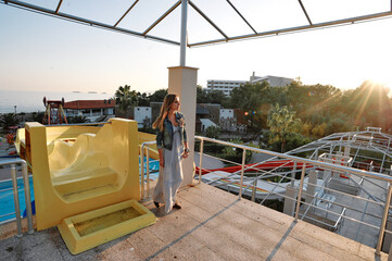 Fototapeta premium A young woman stands on a resort terrace beside a yellow waterpark slide, gazing toward the sea as the sun sets, with pools, palm trees and hotel buildings visible in the warm evening light.