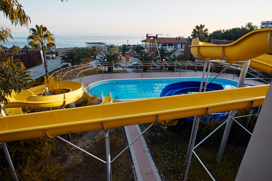 An empty seaside waterpark featuring bright yellow waterslides spiraling above a blue pool at a coastal resort, palm trees and ocean views creating a quiet summer vacation atmosphere.
