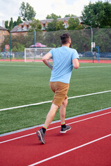 Obraz premium A man in casual attire jogging along a red athletic track in an outdoor sports environment with a green grass field and residential buildings in the background.