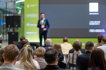 Fototapeta premium Speaker addresses a seated audience beside a dark presentation slide on a large screen.