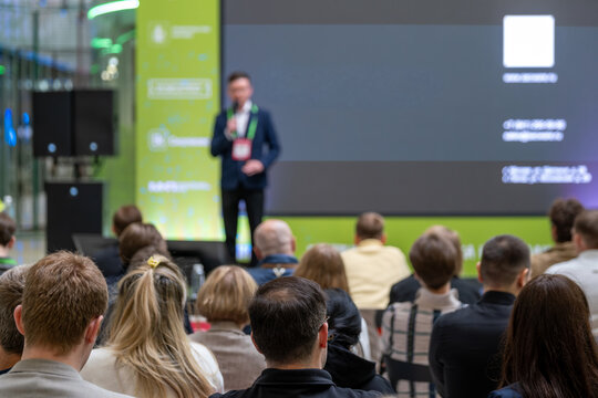 Speaker addresses a seated audience beside a dark presentation slide on a large screen.