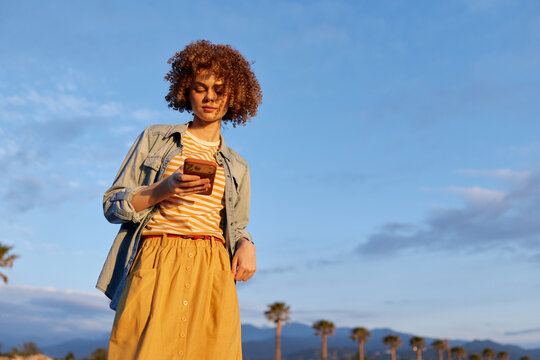 Young woman with curly hair stands outdoors in sunny weather, looking at her smartphone, wearing a striped top, denim jacket and mustard skirt, palm trees line the background.