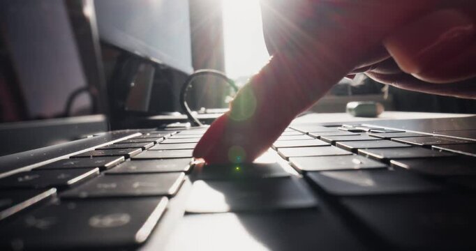 Handheld close up of fingers typing on laptop keyboard at sunlit desk near window, low angle tracking shot.