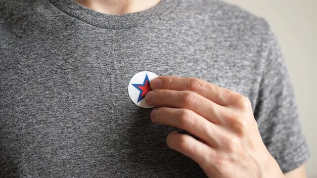A person carefully attaches a circular sticker featuring a red star onto their grey t-shirt, smooths it firmly into place, and then pulls their hand away to show the final result.