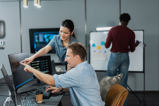 Man developer points at code on a monitor while a woman colleague leans in to review it, with a third teammate writing on a whiteboard in a modern office.