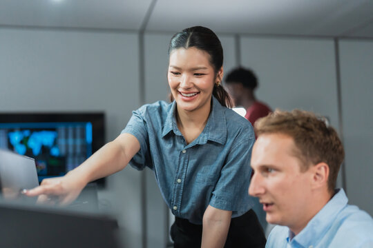 Smiling woman developer points at a monitor beside a male colleague, indicating progress on a software project, with a teammate working behind them in a tech office.