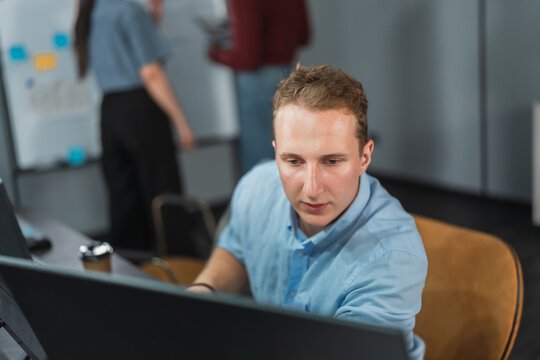 Young programmer concentrates on his screen at a desk while colleagues discuss ideas at a whiteboard in the background of a tech office.