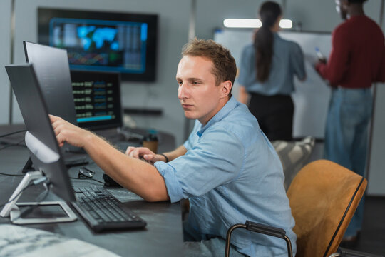 Man developer reaches toward his monitor while debugging an AI model, data dashboards on nearby screens and colleagues planning at a whiteboard behind him.