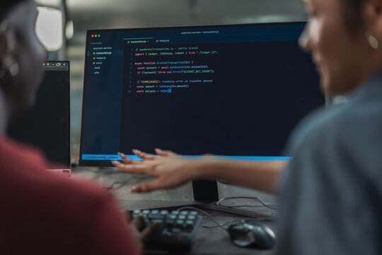 Two colleagues review a screen of program code together at a workstation, with a hand gesturing toward the display in a dark office.
