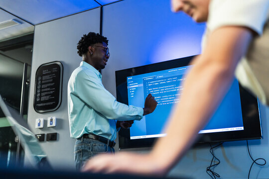 Male developer gestures toward lines of code displayed on a wall screen during a technical discussion in a blue-lit office.