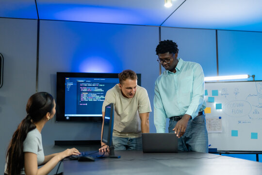 Two colleagues lean over a laptop to review work while a teammate types at a desktop in a blue-lit software development office.